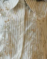 Camisa de corte clásico a rayas verticales en tonos neutros, con cuello camisero y cierre frontal de botones. Presenta un delicado bordado floral con canutillos y lentejuelas brillantes en el frente.