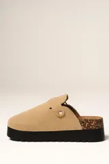 Zueco de plataforma color beige, con capellada de gamuza sintética, detalle de hebilla metálica lateral y suela de goma negra con detalle de corcho en el talón.