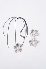 Set compuesto por un collar con cordón negro y un par de aros, ambos con dijes en forma de flor de metal plateado con textura irregular y una perla central.
