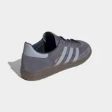 Championes Adidas Handball Spezial de gamuza color gris con las icónicas tres tiras laterales en un tono más claro y suela de caucho natural. Presenta el logo de la marca y el nombre del modelo en dorado en el lateral.