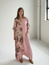Vestido largo color rosa, con mangas cortas y diseño holgado, presenta un estampado floral en tonos marrones y verdes en un lateral, además de un bolsillo aplicado en la parte frontal y costuras visibles de estilo rústico.