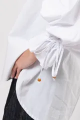 Camisa de algodón blanca de silueta amplia, con cuello camisero, botonadura frontal con botones decorativos, mangas abullonadas extragrandes y detalles de tiritas ajustables con cuentas de resina en los puños.