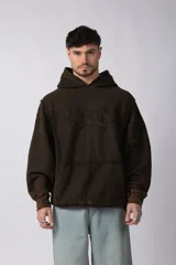 Buzo canguro oversize de algodón con efecto de lavado gastado, color marrón oscuro. Presenta un aplique decorativo cosido en el pecho con diseño abstracto y costuras expuestas.