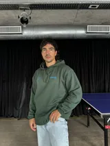 Canguro verde oliva con capucha y cordones ajustables. Estampado en la espalda con la frase "Bad Ideas Good Stories" en letras azules.