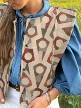 Chaleco de tela estampada con diseño geométrico en tonos beige, bordó y gris, con hombreras estructuradas y cierre frontal con broches a presión metálicos.