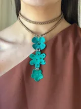 Choker de doble vuelta con tiento de gamuza trenzado en color marrón y dije colgante compuesto por tres flores de metal texturizado en color turquesa con acabado brillante y detalles de mostacillas.