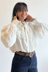 Blusa blanca de estilo romántico con volados tipo shabot en el frente, cuello alto con lazo ajustable y mangas largas abullonadas con puños ajustados.