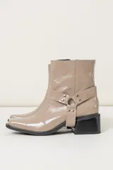 Botas cortas color visón de cuero charolado, con punta cuadrada, caña media y detalle de arnés con hebillas plateadas. Taco cuadrado de altura media color negro.