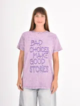 Remera de algodón con efecto acid wash en color lila, de corte holgado y manga corta. Presenta un estampado frontal con la frase 'Bad choices make good stories' en letras grandes de color violeta.