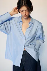 Camisa de manga larga con cuello solapa y hombreras. Presenta un diseño de rayas verticales en tonos azul y blanco, con cierre frontal de botones y puños anchos.