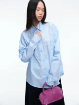Camisa de corte oversize color celeste, con cuello clásico, cierre frontal de botones y detalle de apliques brillantes en la parte delantera.