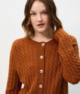 Cárdigan de punto trenzado color marrón, con cuello redondo, manga larga y cierre frontal con botones metálicos. Presenta puños y ruedo acanalados.