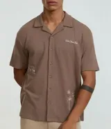Camisa de manga corta, color marrón topo, con cuello camisero y corte amplio (boxy). Presenta bordados minimalistas en blanco en el frente, simulando un paisaje.