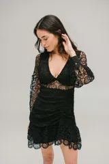 Vestido corto de encaje negro con diseño calado, escote en V, mangas largas con puños y falda con volados. Presenta un corte ajustado con detalle fruncido en la cintura y cierre invisible.