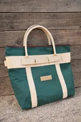 Bolso de playa de lona color verde oscuro con detalles en cuero beige y marrón. Tiene asas de cuero beige y una correa decorativa en la parte superior con cierre de botón. Incluye un parche con el logo de la marca "Sierra Mora".
