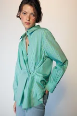Camisa de manga larga con cuello solapa, cierre frontal con botones y diseño cruzado con lazada lateral en el mismo tejido. Presenta un estampado de rayas verticales en tonos verdes y azules.
