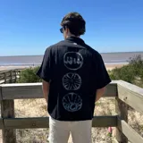 Camisa de lino negra con corte boxy, manga corta y cuello camisero. Presenta un estampado gráfico en la espalda compuesto por tres círculos alineados verticalmente.