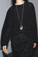 Blusa de cuello redondo y manga larga, confeccionada en tejido fluido con acabado satinado. Presenta un diseño de corte holgado, bajo recto y cierre en la espalda mediante una pequeña abertura con botón.