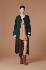 Saco Natalia Antolin de mujer color verde oscuro, de corte largo y diseño relajado con cuello redondo, mangas anchas y cierre frontal de botones oscuros. Incluye una etiqueta decorativa distintiva de la marca en el puño de la manga izquierda.