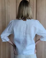 Camisa de manga tres cuartos color beige con cuello con volados y botones frontales. Presenta un detalle de color negro en la parte superior de las mangas.