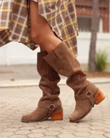 Botas de caña alta confeccionadas en cuero gamuzado color marrón. Presentan un diseño de estilo western con puntera cuadrada, taco de madera y una tira decorativa con estribo metálico desmontable en la zona del tobillo.