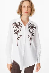 Camisa blanca de corte clásico con cuello camisero y cierre frontal de botones. Presenta un bordado lineal en color marrón oscuro en ambos lados del frente, representando motivos florales.
