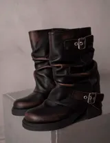 Botas de caña media con efecto desgastado en tono marrón oscuro. Presentan un diseño holgado con pliegues, dos correas ajustables con hebillas metálicas y taco cuadrado bajo.