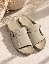 Sandalias planas color beige, fabricadas en gamuza, con tira ancha en el empeine, hebilla metálica de ajuste y detalle de flecos colgantes con argolla.
