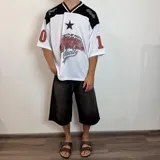 Camiseta estilo fútbol americano de corte oversize, color blanco con detalles en negro y rojo. Presenta estampado gráfico frontal con estrella y texto, además de números en las mangas.