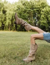 Bota alta de gamuza color beige claro, estilo cowboy o western, con caña arrugada y taco bajo y ancho.