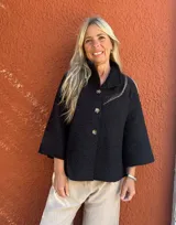 Capa corta de textura tipo bucle en color negro, con cuello alto, cierre frontal de botones y mangas tres cuartos con corte raglán y silueta en A.