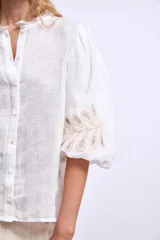 Blusa blanca de lino con cuello mao, mangas 3/4 abullonadas con apliques bordados en forma de hojas y cierre frontal con botones.
