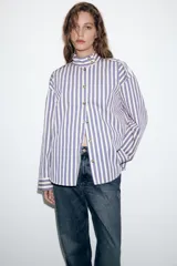 Camisa de manga larga con cuello subido y trabillas en los hombros. Presenta un diseño de rayas verticales en tonos azul, beige y marrón, con cierre frontal de botones.