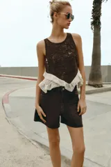 Top sin mangas con cuello redondo, confeccionado en una combinación de tejido de crochet marrón con diseño floral y base de tela color crudo con volados asimétricos y detalles de puntilla. Cuenta con cierre de botón en la espalda.