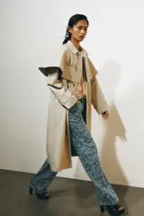 Gabardina larga de corte recto y diseño minimalista en color beige, con cuello solapa, manga larga y cierre frontal con botones. Cuenta con una capa desmontable en el mismo tejido y una solapa decorativa en el pecho.