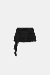 Falda pantalón de tiro medio color negro, con diseño de volantes y tejido asimétrico lateral que incluye un lazo colgante. Cuenta con cierre lateral de cremallera oculta.
