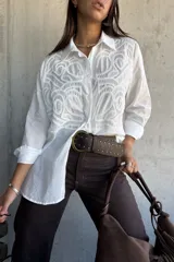 Camisa blanca de corte oversize con diseño de bordado calado en el frente. Presenta cuello clásico, cierre frontal con botones y mangas largas.