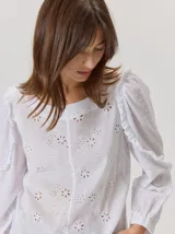 Camisa blanca de algodón con diseño de bordado inglés, cuello redondo, mangas largas con volados en los hombros y puños elásticos.