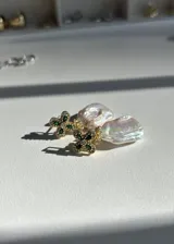 Aros colgantes con forma de trébol de cuatro hojas con circonitas verdes y perla barroca de río. El metal es plata 925 con baño de oro.