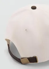 Gorro tipo gorra con visera, confeccionado en algodón, con diseño bicolor en color crudo y marrón, cierre regulable con hebilla metálica y detalle de mensaje bordado.
