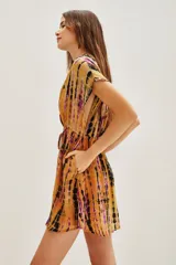 Vestido corto de estilo kimono con estampado tie-dye en tonos mostaza, violeta y negro. Presenta escote en V profundo, mangas cortas amplias y ajuste en la cintura con cordón. Incluye detalles de bordados decorativos con cuentas en el escote y la falda.