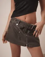 Minifalda de denim gris con efecto lavado, corte de tiro bajo y cierre frontal con cremallera. Incluye un cinturón integrado con ojales metálicos y un bolsillo frontal con cierre.