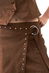 Minifalda de gamuza símil color marrón, con diseño envolvente tipo wrap, detalle de tachas metálicas en el frente y cinturón integrado con aro metálico.