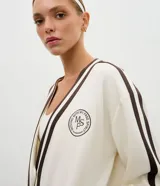 Cardigan deportivo de corte oversized, color blanco con detalles en marrón oscuro. Presenta escote en V, cierre frontal con botones a presión, mangas largas con franjas laterales y puños acanalados. Incluye un parche circular con letras en el pecho.