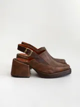 Zapato tipo zueco de cuero color chocolate con efecto desgastado, plataforma y taco cuadrado. Presenta una tira trasera ajustable con hebilla metálica.