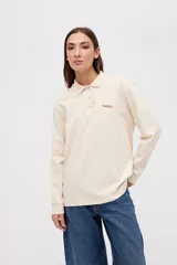 Remera tipo polo de manga larga en color beige, confeccionada en algodón pesado con textura suave. Presenta cuello clásico, cierre frontal con botones y un pequeño bordado en el pecho. Posee puños en las mangas y un corte relajado.