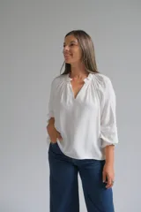 Blusa blanca de corte holgado con cuello redondo fruncido y abertura frontal en V. Presenta mangas tres cuartos con puños elásticos y detalle de tejido texturizado en los mismos.