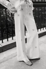 Pantalón de jean blanco, tiro superalto, con piernas amplias y corte holgado. Presenta tachas decorativas en forma de estrella o copo de nieve distribuidas por toda la prenda.