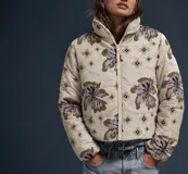 Campera puffer color beige con estampado floral y de logotipos en tonos marrones y grises, cuello alto y cierre frontal con cremallera.