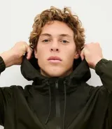 Campera rompevientos de color gris oscuro, con capucha ajustable mediante cordones, cierre frontal por cremallera y bolsillos laterales con cierre. Presenta puños elásticos y un diseño liso y minimalista.